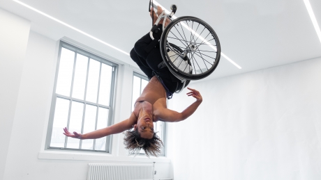 Alice Sheppard hovers upside down, suspended from the ceiling in her wheelchair, in a brown leotard and black leggings. She is a light-skinned, multiracial Black woman with short curly hair. Her arms extend as if caught mid-flight. Photo by Mengwen Cao.