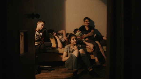 The Earth is Blue as an Orange Looking through the an open door into a room, five children sit on a coach all looking at the opposite wall and smiling.