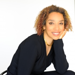 Alice Sheppard hovers upside down, suspended from the ceiling in her wheelchair, wearing a brown leotard and black leggings. Alice Sheppard, a multi-racial Black woman with coffee-colored skin, blond, copper, and red striped curly hair gazes toward the camera. She wears a black shirt; her face rests in her hand, and a gold necklace gleams at her neck. Photo by Beverlie Lord.