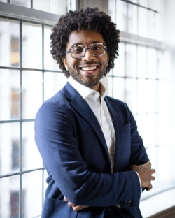 Seth Parker Woods stands in front of a window smiling, with his arms crossed, wearing a dark blue suit and black and white speckled round rimmed glasses.